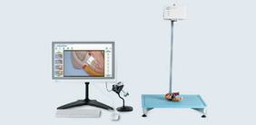 Image displays a medical simulation setup, featuring a monitor showing anatomical visuals, a camera on a stand, and a blue platform with a model figure on it. This arrangement is likely used for educational or training purposes in medical fields.