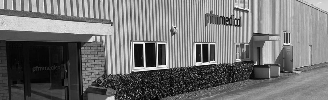 The image shows the exterior of a medical facility building featuring a modern design. The entrance is highlighted, with the name "pfm medical" prominently displayed on the wall. The building has vertical metal siding and several windows, surrounded by green foliage.