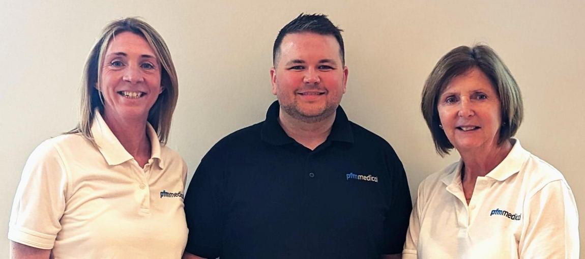 Three individuals are standing close together, smiling. Two women are wearing white polo shirts with a logo, and a man in the center is wearing a black polo shirt with a logo. They appear friendly and are against a plain background.