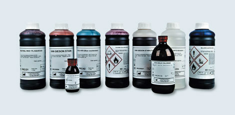 A row of various bottles containing liquids, each labeled with safety symbols and descriptions. The bottles differ in size and color, including shades of red, blue, and purple, indicating different chemical contents. Some bottles have warning labels for hazardous materials.
