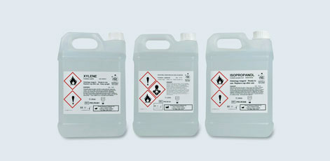 Three clear plastic containers labeled with hazardous material warnings. The labels indicate the contents as Xylene and Isopropanol, displaying symbols for flammability and health hazards. Each container has a handle and a secure screw cap.